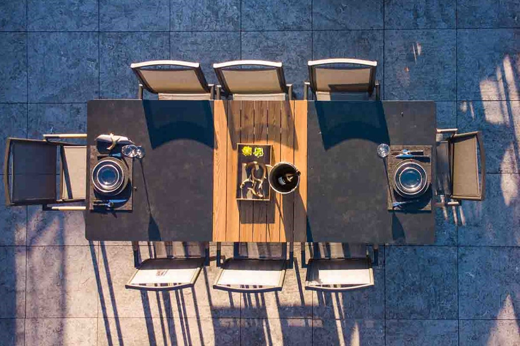 A bird's-eye view of a black outdoor dining table with a ceramic glass & brown wooden pattern tabletop design. Surrounding the table are 8 outdoor aluminium chairs, forming a set. The furniture is on stone tiles in an outdoor area.