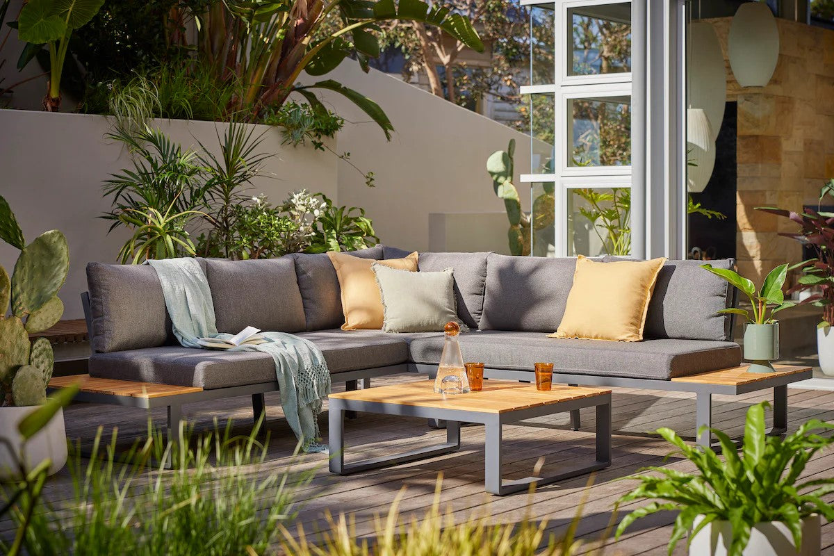 An outdoor patio featuring an aluminium outdoor lounge with grey cushions and yellow pillows, on a wooden deck. There is a wooden and aluminium coffee table with two glasses of water and a flask of water on it. The area is surrounded by abundant greenery.