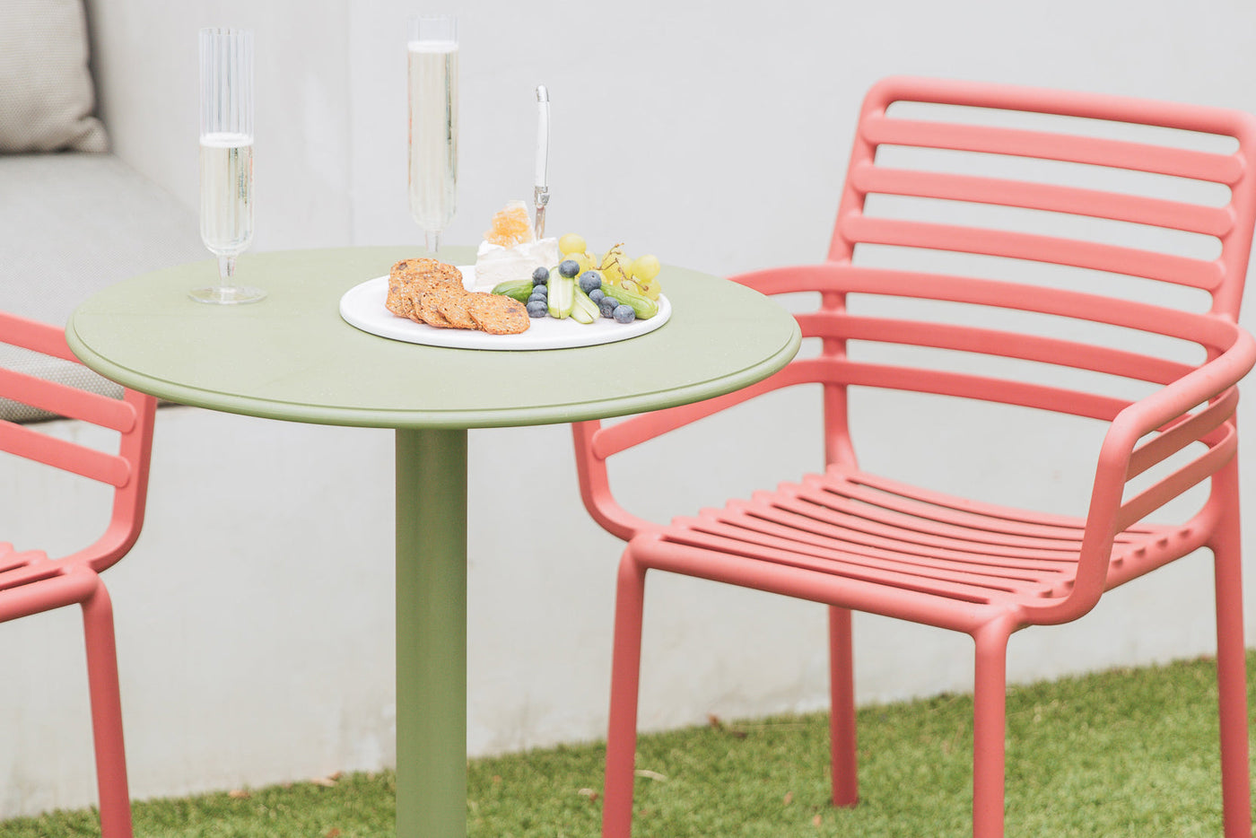 Green outdoor Nardi balcony table set with fruit, crackers, and wine, paired with two salmon pink Nardi chairs in a cozy backyard setting.