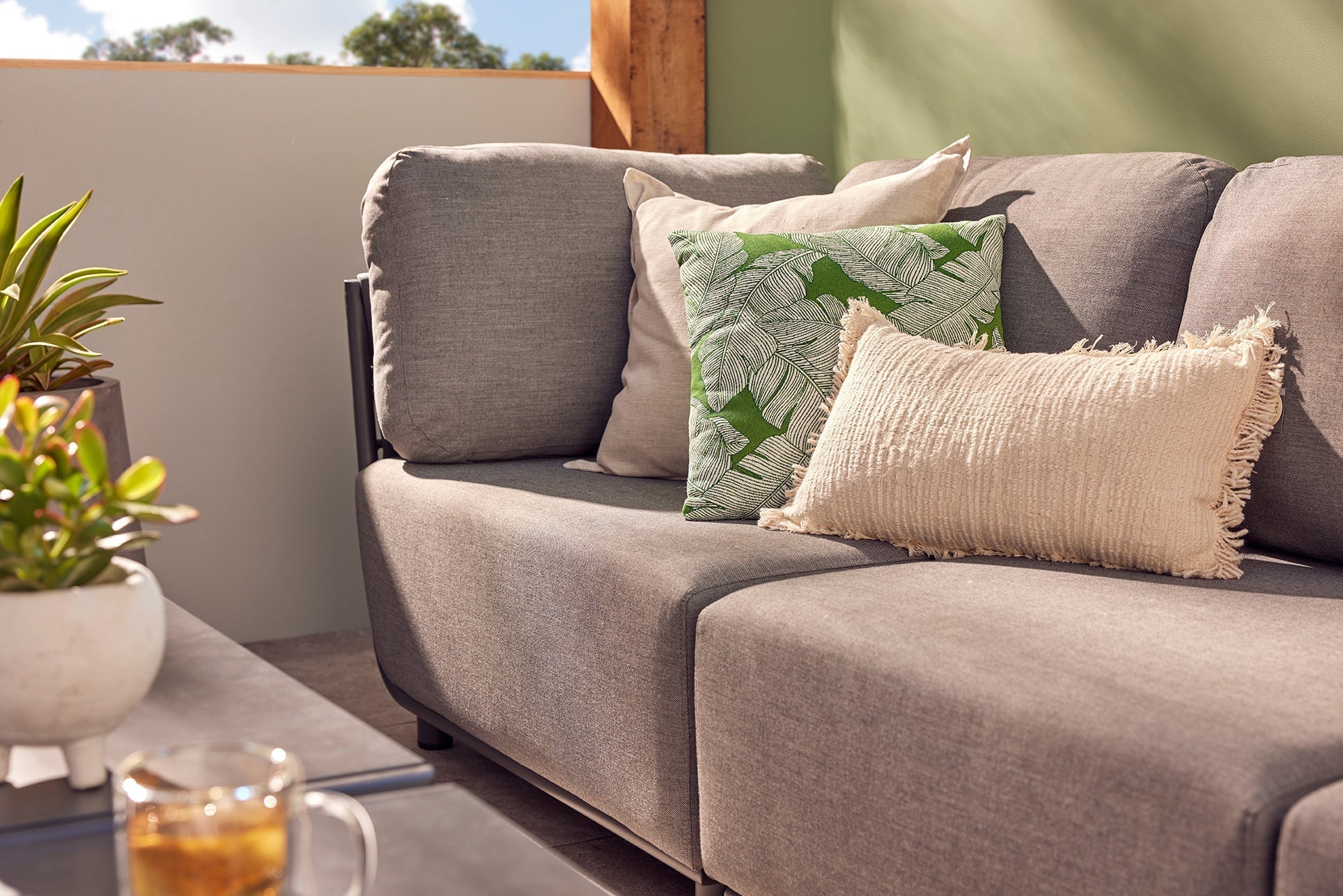 A modern grey outdoor lounge with 3 decorative pillows and an outdoor coffee table, set against a green wall. We see 2 pot plants & a cup of tea on the coffee table.