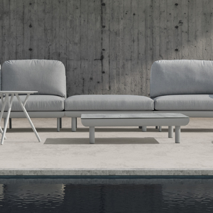 A modern large outdoor sofa set in light grey with an ottoman is shown in a close-up shot of a modern concrete industrial outdoor setting.