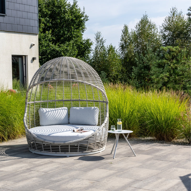 The Peninsula Outdoor Wicker Daybed by Couture Jardin sits on a tiled patio surrounded by lush greenery. Featuring a dome-shaped wicker design with light grey cushions, it’s paired with a white side table holding lemonade and a book. Shadows from the open weave add elegance, with a modern house visible in the background.