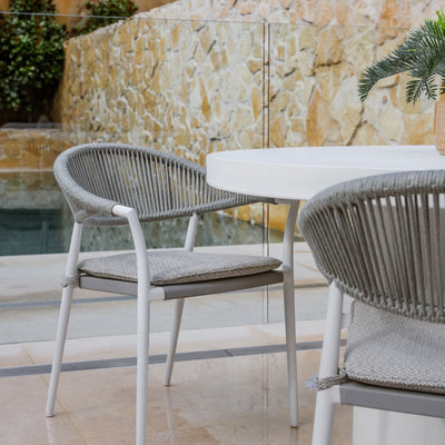 A modern outdoor dining rope chair with a sleek, minimalist design. It features a curved backrest with evenly spaced vertical spindles and a simple, flat seat. The chair is made of white aluminium and is set against an outdoor backdrop with greenery.