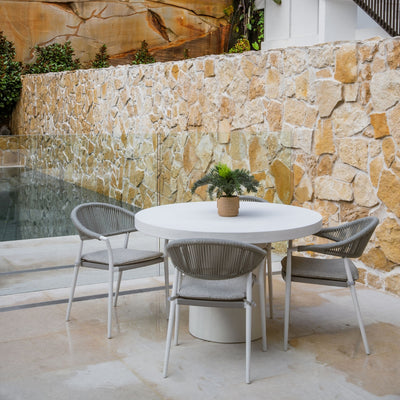 Four contemporary outdoor dining rope chairs with sleek, minimalist designs, paired with a white round outdoor dining table. The outdoor furniture is arranged in a backyard with a pool, some rocks, and greenery.