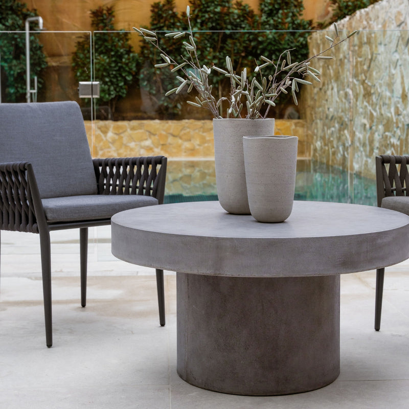 Zen round outdoor concrete coffee table, 90 cm, with a dark-grey weathered cement colour, in an outdoor setting with two outdoor armchairs. The area is by a modern-looking pool with lots of sandstone in the background.