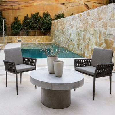 Zen round outdoor concrete coffee table, 90 cm, with a dark-grey weathered cement colour, in an outdoor setting with two outdoor armchairs. The area is by a modern-looking pool with lots of sandstone in the background.