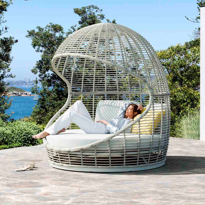 A lady enjoying the sunshine on a wicker daybed by the river, surrounded by greenery for privacy, capturing the essence of outdoor living.